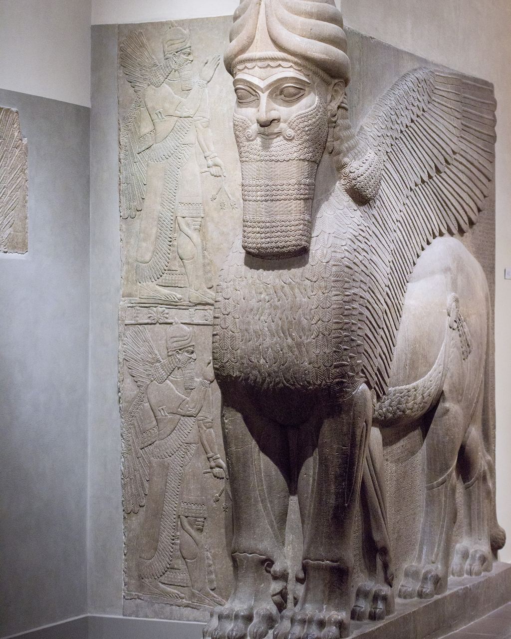 Assyrian lamassu, a colossal human-headed winged lion carved in stone, standing guard in a museum gallery with a palace relief on the wall behind