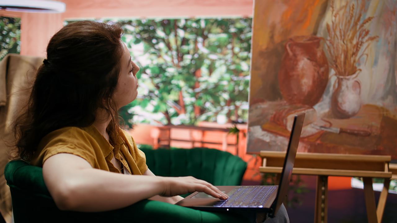 A woman browsing images of artworks on a laptop in a studio, with paintings on the wall behind