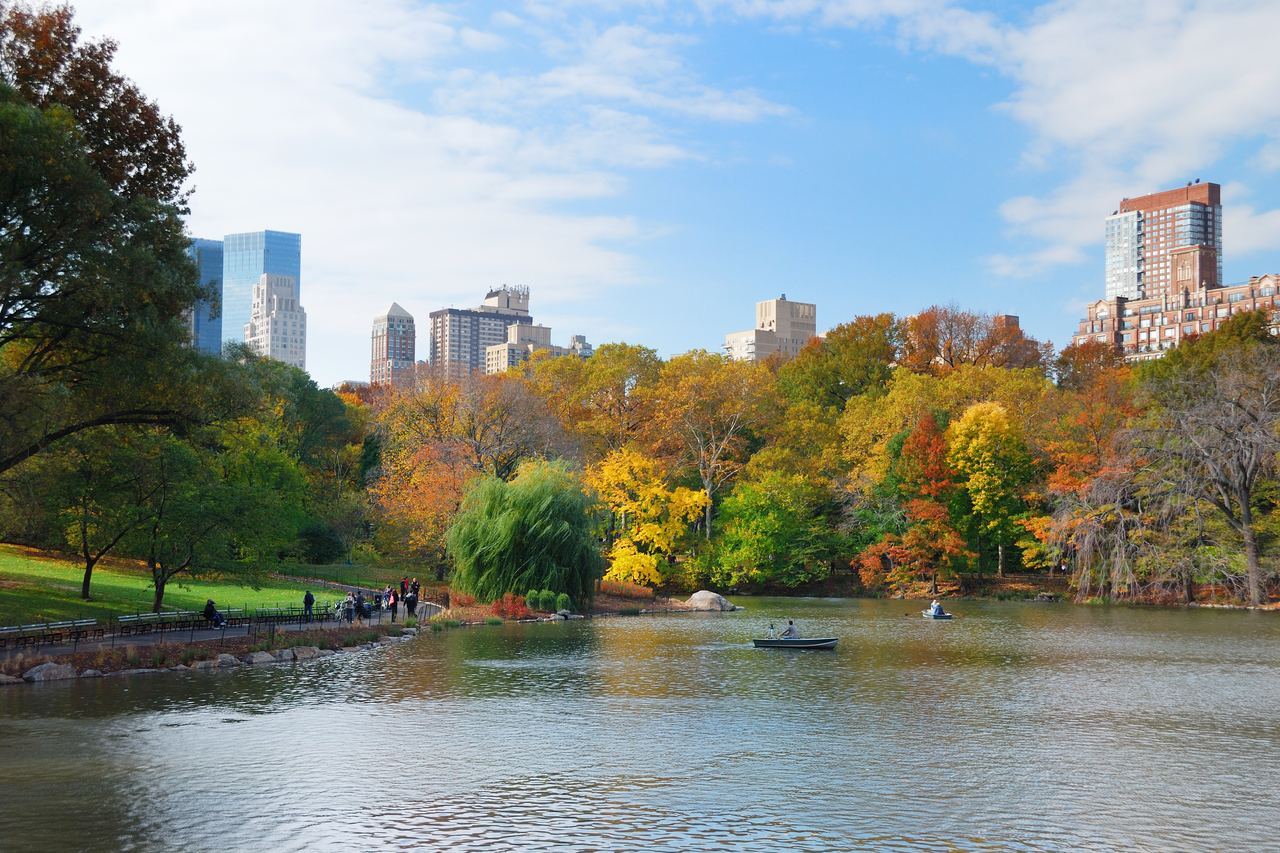 The Lake in Central Park in autumn, with red and yellow trees along the shore, rowboats on the water and Manhattan towers rising above the foliage