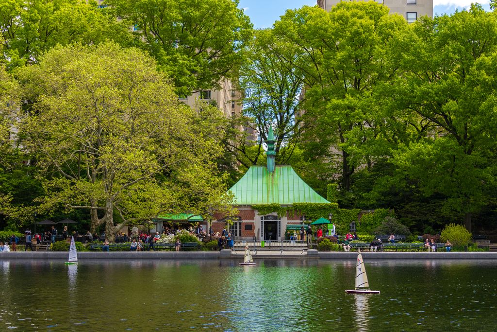 A model sailboat on Conservatory Water in front of the green-roofed Kerbs Boathouse in Central Park, New York