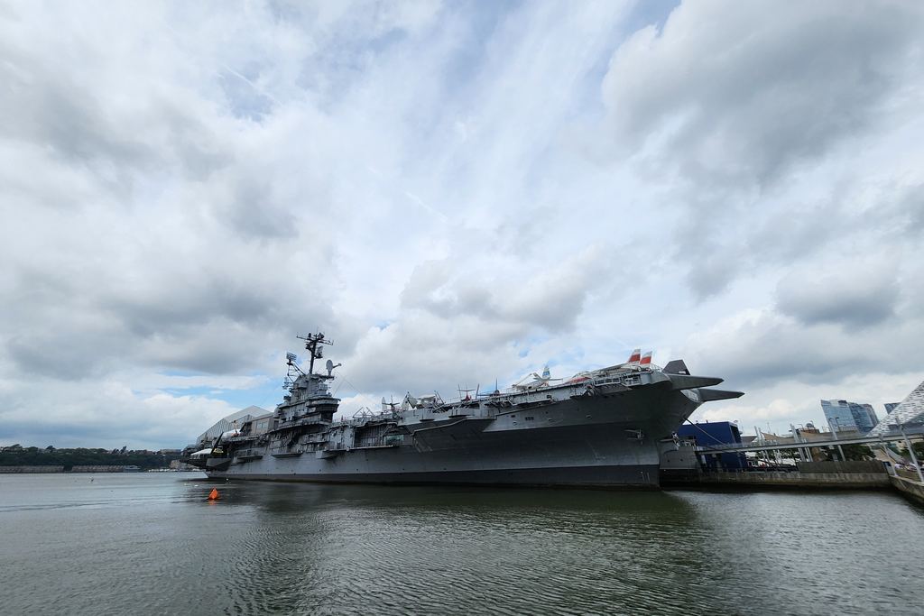 Wide view across the Hudson River of the aircraft carrier Intrepid at its Manhattan pier, now the Intrepid Museum, under a dramatic cloudy sky