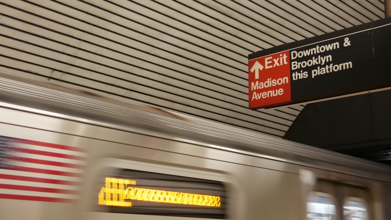 A New York City subway train pulling into an underground platform, with a sign pointing to the Madison Avenue exit