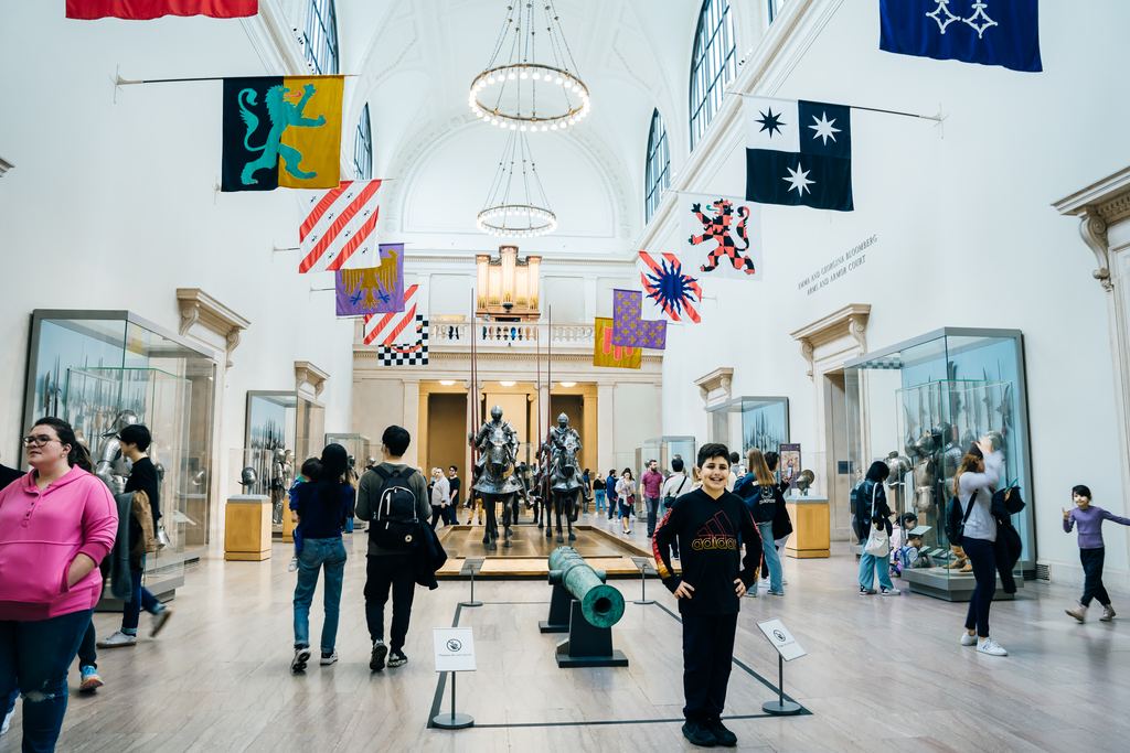 The Arms and Armor court of the Metropolitan Museum of Art, with heraldic banners hanging from the vault, mounted armour and visitors among the display cases