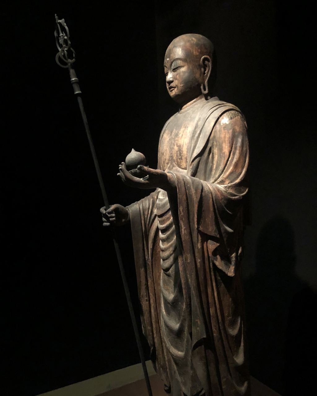 A carved wooden statue of a Buddhist monk holding a staff, in the Asian art galleries of the Metropolitan Museum of Art