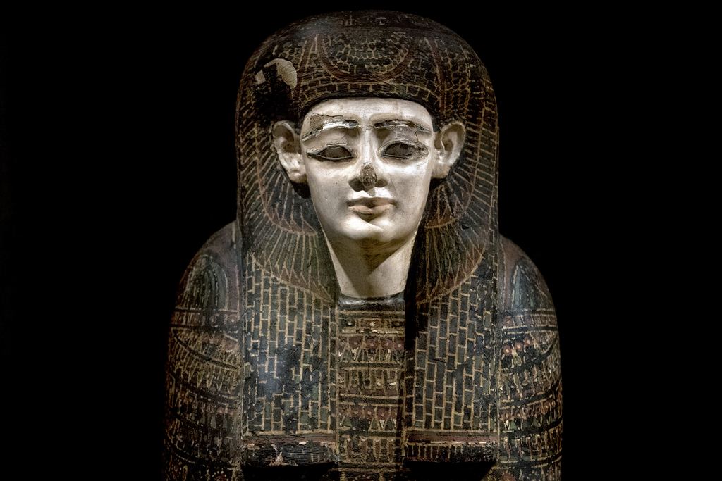 The pale painted face of an ancient Egyptian coffin lit against a dark background at the Metropolitan Museum, New York