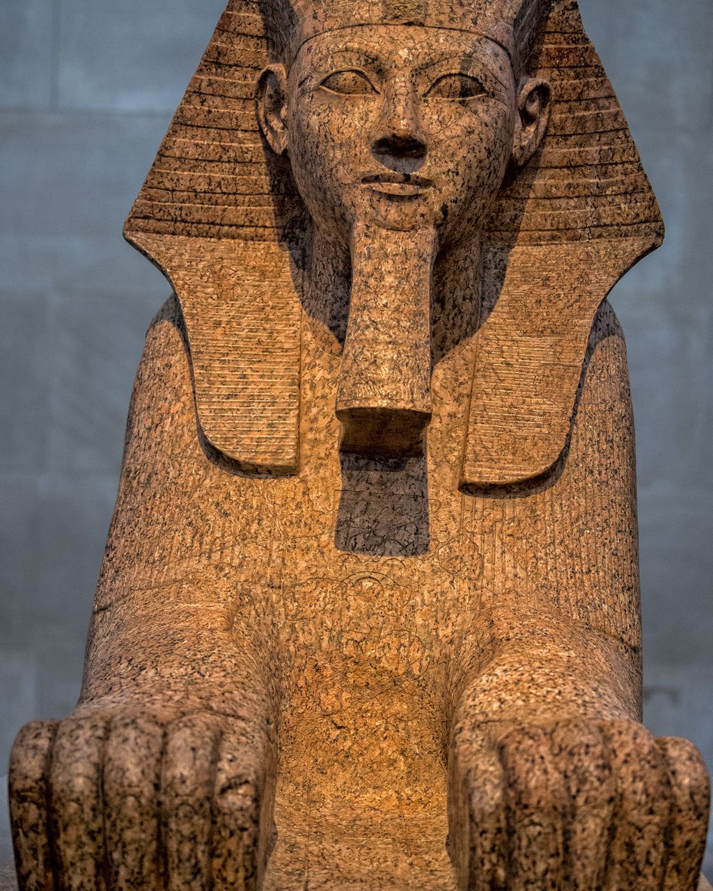 A red granite sphinx with a pharaoh's striped headdress and lion's paws, displayed in the Egyptian galleries of the Metropolitan Museum of Art