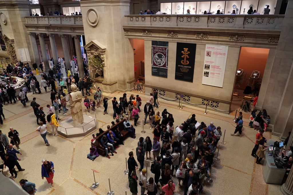 A busy Great Hall at the Metropolitan Museum of Art seen from the balcony, with visitors gathered around the central desk and exhibition banners on the wall