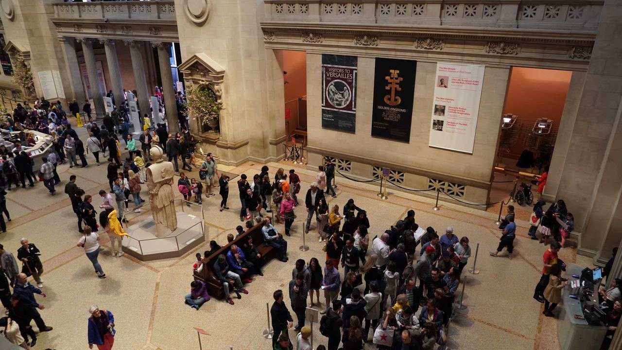 A busy Great Hall at the Metropolitan Museum of Art seen from the balcony, with visitors gathered around the central desk and exhibition banners on the wall