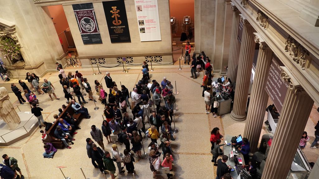 Visitors queuing at the desks in the Great Hall of the Metropolitan Museum of Art, seen from the balcony above the entrance floor