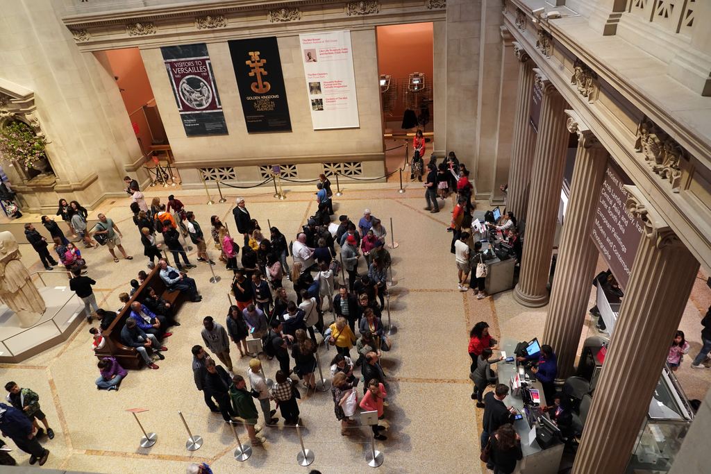 Visitors queuing at the desks in the Great Hall of the Metropolitan Museum of Art, seen from the balcony above the entrance floor