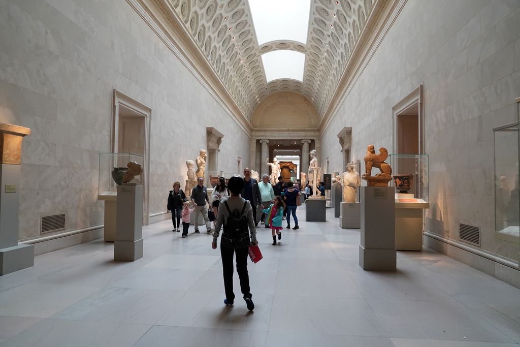 The long barrel-vaulted hall of Greek sculpture at the Metropolitan Museum of Art, with marble statues and vases on pedestals and visitors walking down the centre