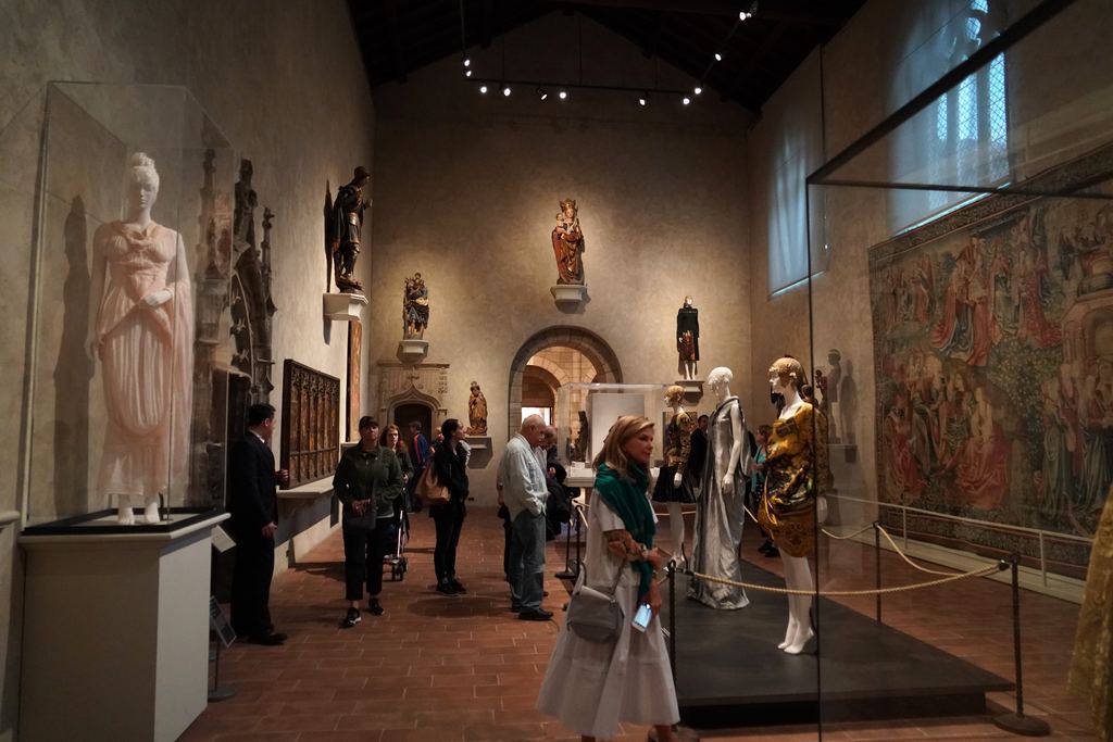 Visitors in a medieval gallery of the Metropolitan Museum of Art during the 2018 Heavenly Bodies exhibition, with statues, a tapestry and fashion on display