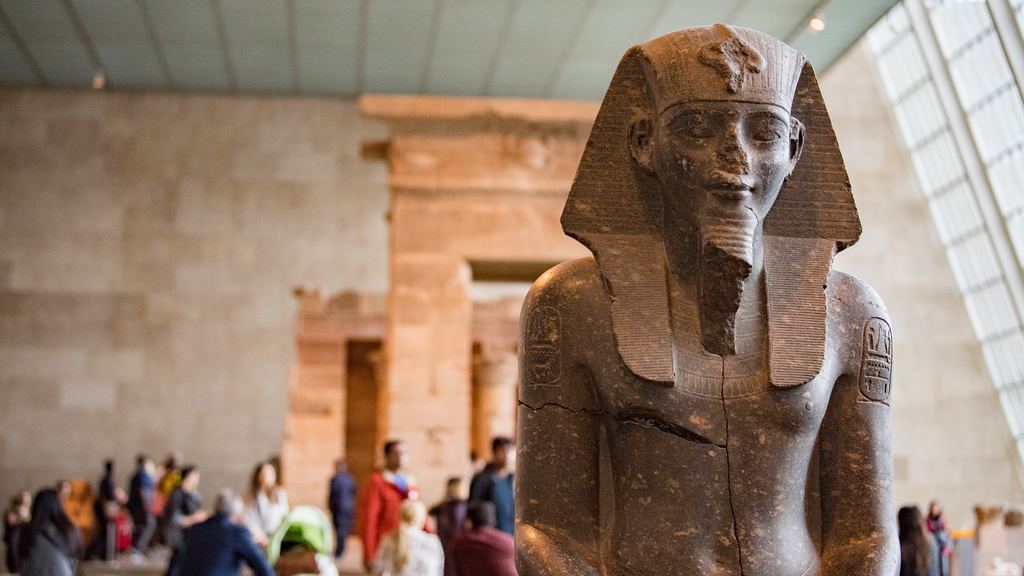 A granite statue of an Egyptian king in gallery 131 of the Metropolitan Museum, with the Temple of Dendur and seated visitors softly out of focus behind