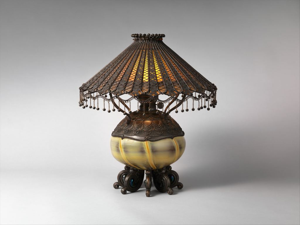 A Tiffany Studios table lamp: a domed bronze filigree shade with a beaded fringe over a glowing amber liner, above a pale banded blown-glass body resting on bronze feet set with blue cabochons