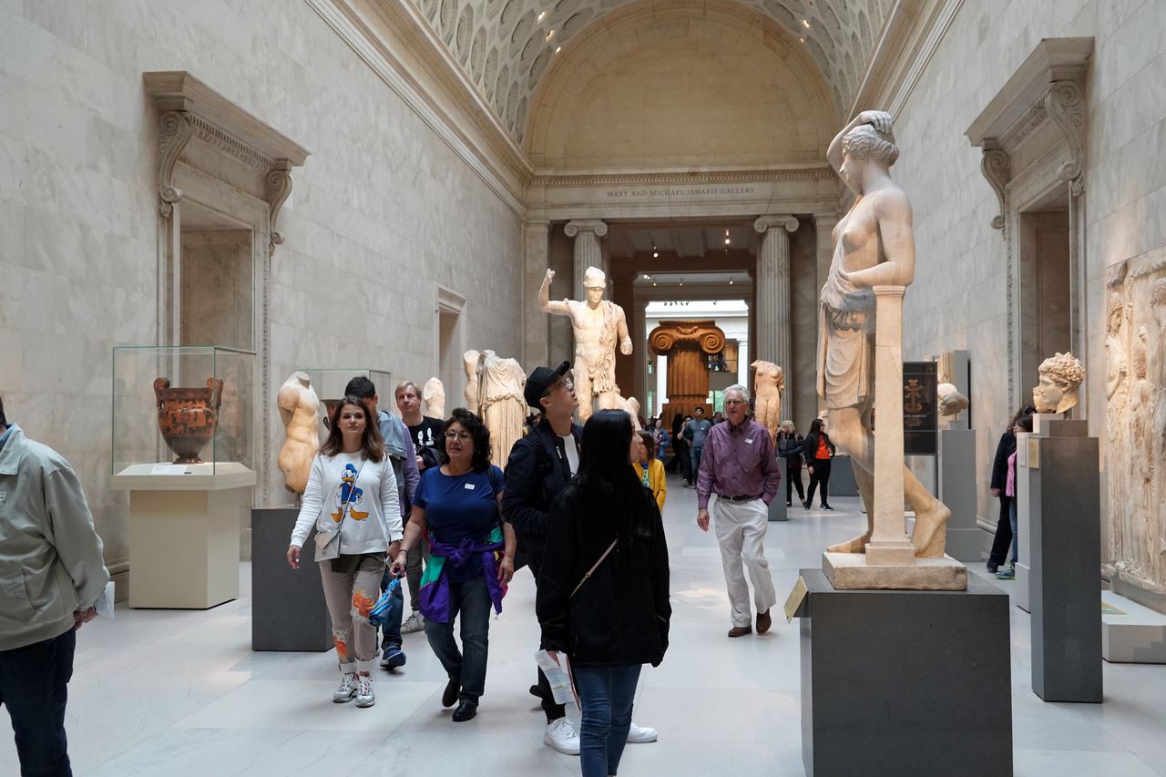 Visitors walking through the vaulted Greek sculpture hall of the Metropolitan Museum of Art, between marble statues and display cases
