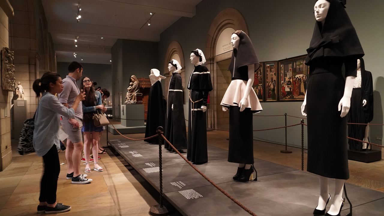Visitors carrying bags walk past a roped-off display of black outfits in a gallery of the Metropolitan Museum of Art, 2018