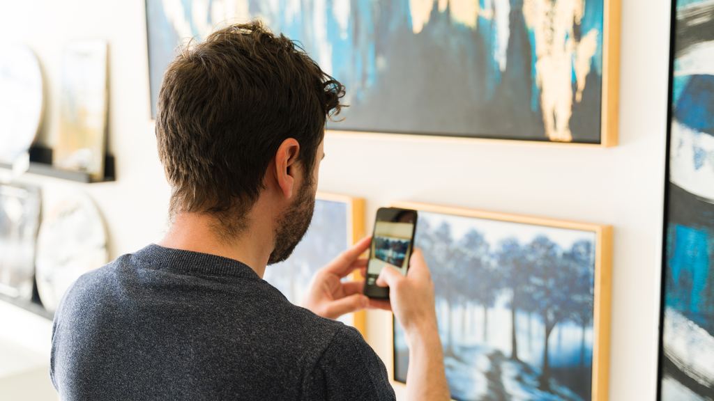A visitor taking a photo of a framed painting with his smartphone in a bright art gallery