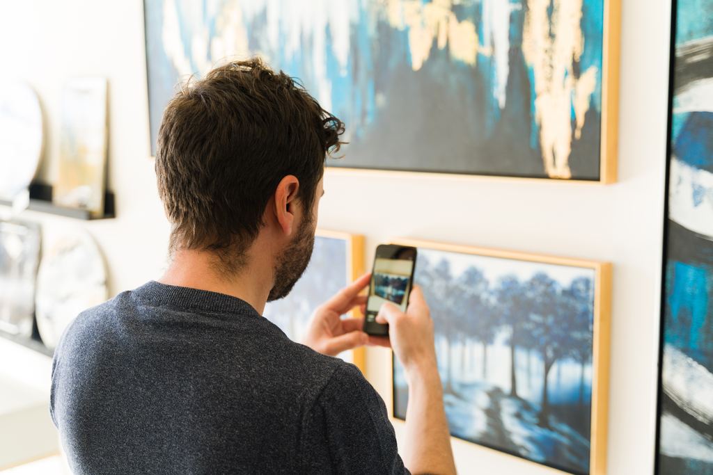A visitor taking a photo of a framed painting with his smartphone in a bright art gallery