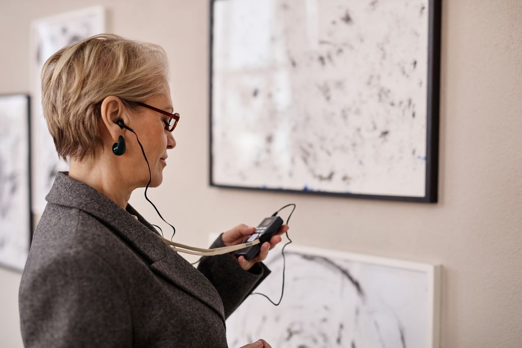 An older visitor listening on earphones to an audio device while looking at drawings in a museum
