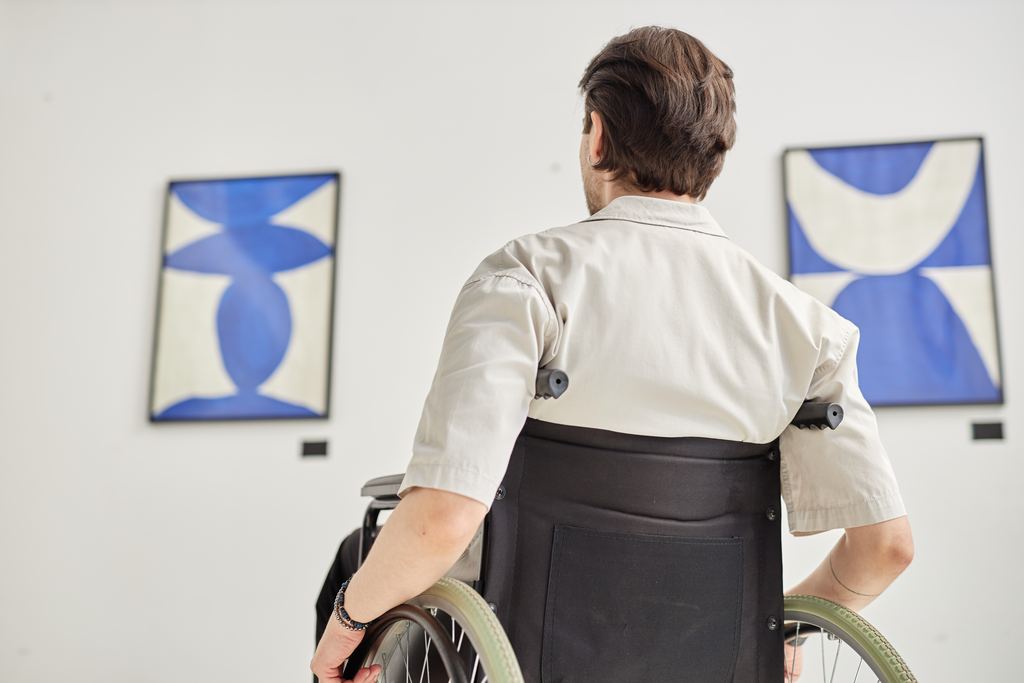 Back view of a wheelchair user looking closely at framed artworks on a gallery wall